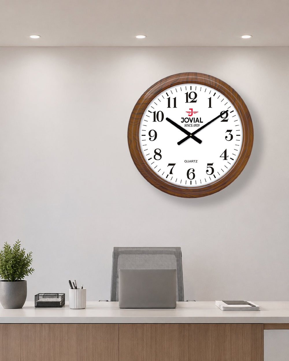 A round wall clock with a brown wooden frame and a white face hangs on a light gray office wall above a desk with a laptop, pen cup, and small plant. The clock shows the brand name 'ZOVIAL' on the face.